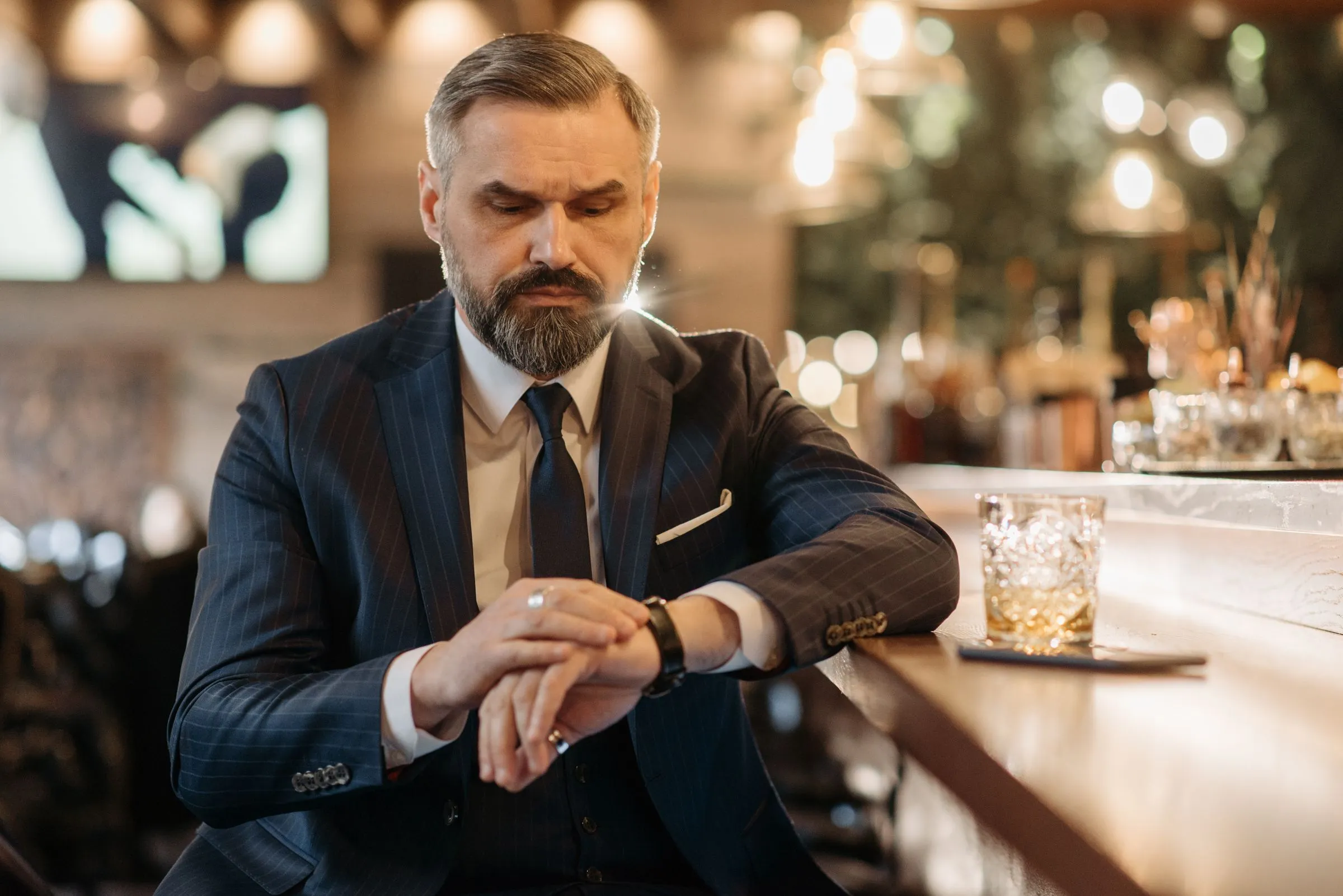 Caballero con traje azul de raya diplomática consultando su reloj en la barra de un bar