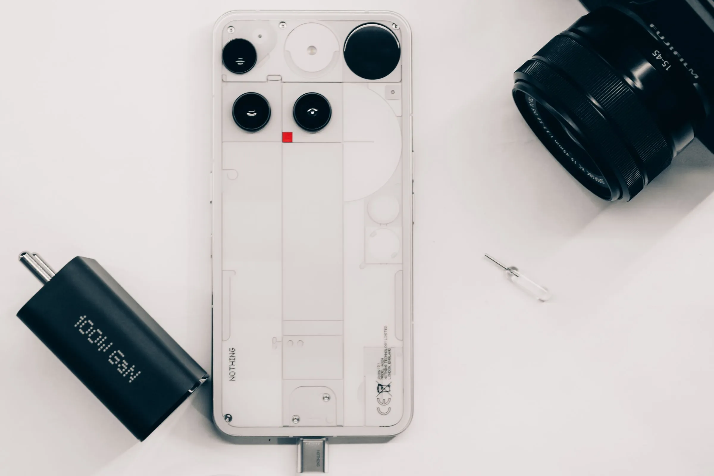 Teléfono de carcasa transparente, objetivo fotográfico y cargador dispuestos sobre fondo blanco
