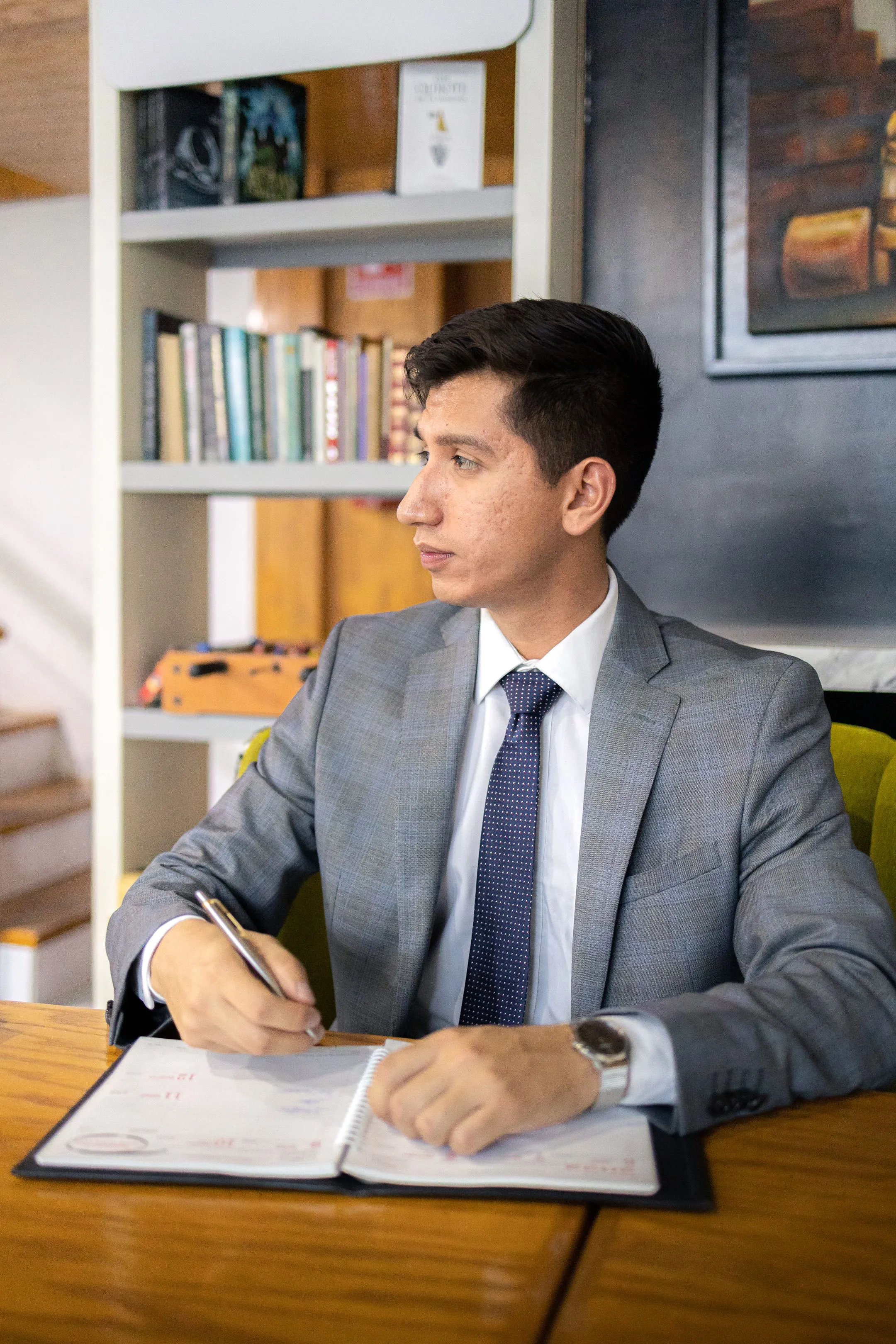 Hombre joven con traje gris y corbata, pluma en mano, sentado a su escritorio ante una estantería de libros