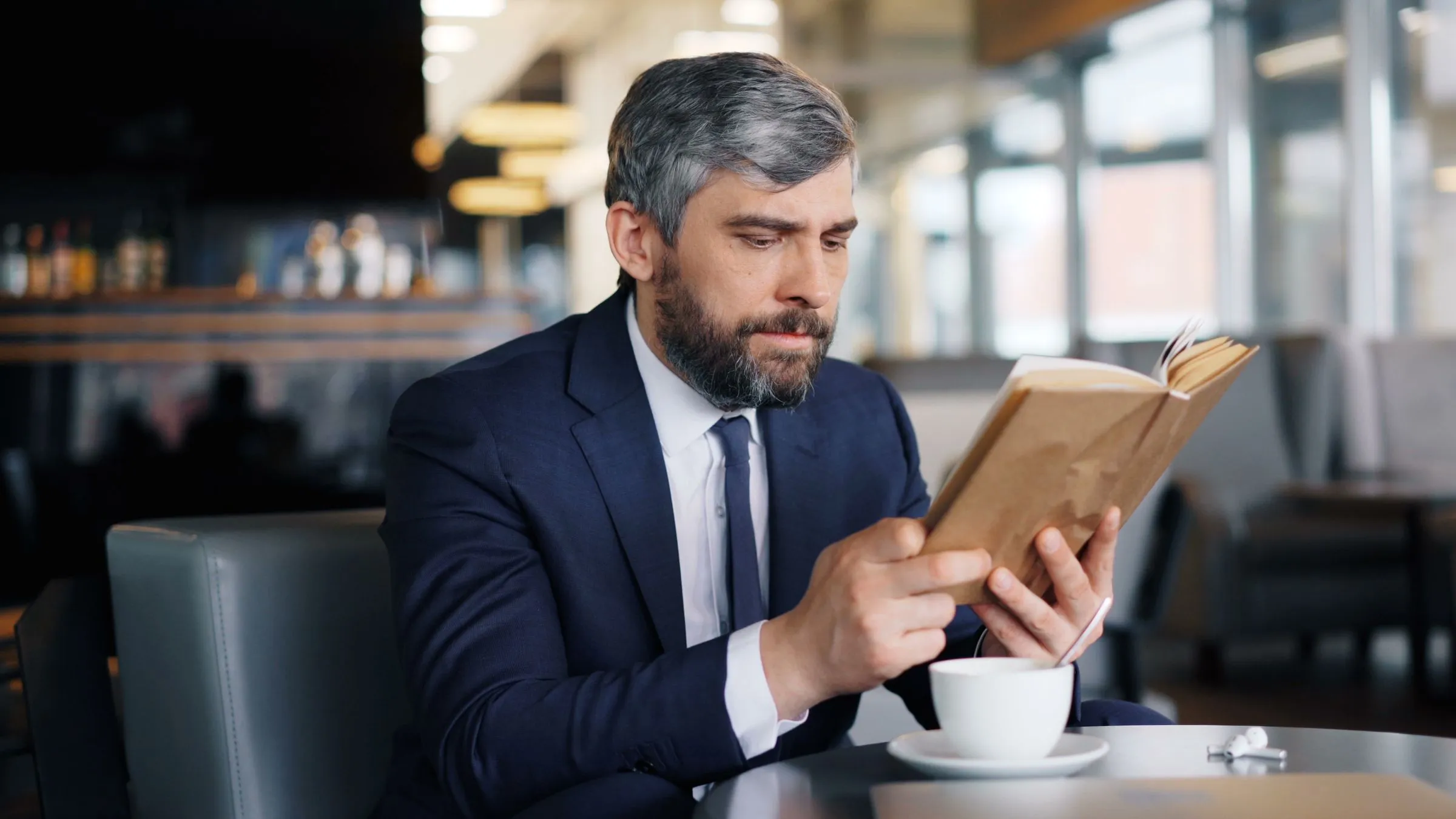Hombre maduro de barba cana y traje azul marino leyendo un libro en una cafetería