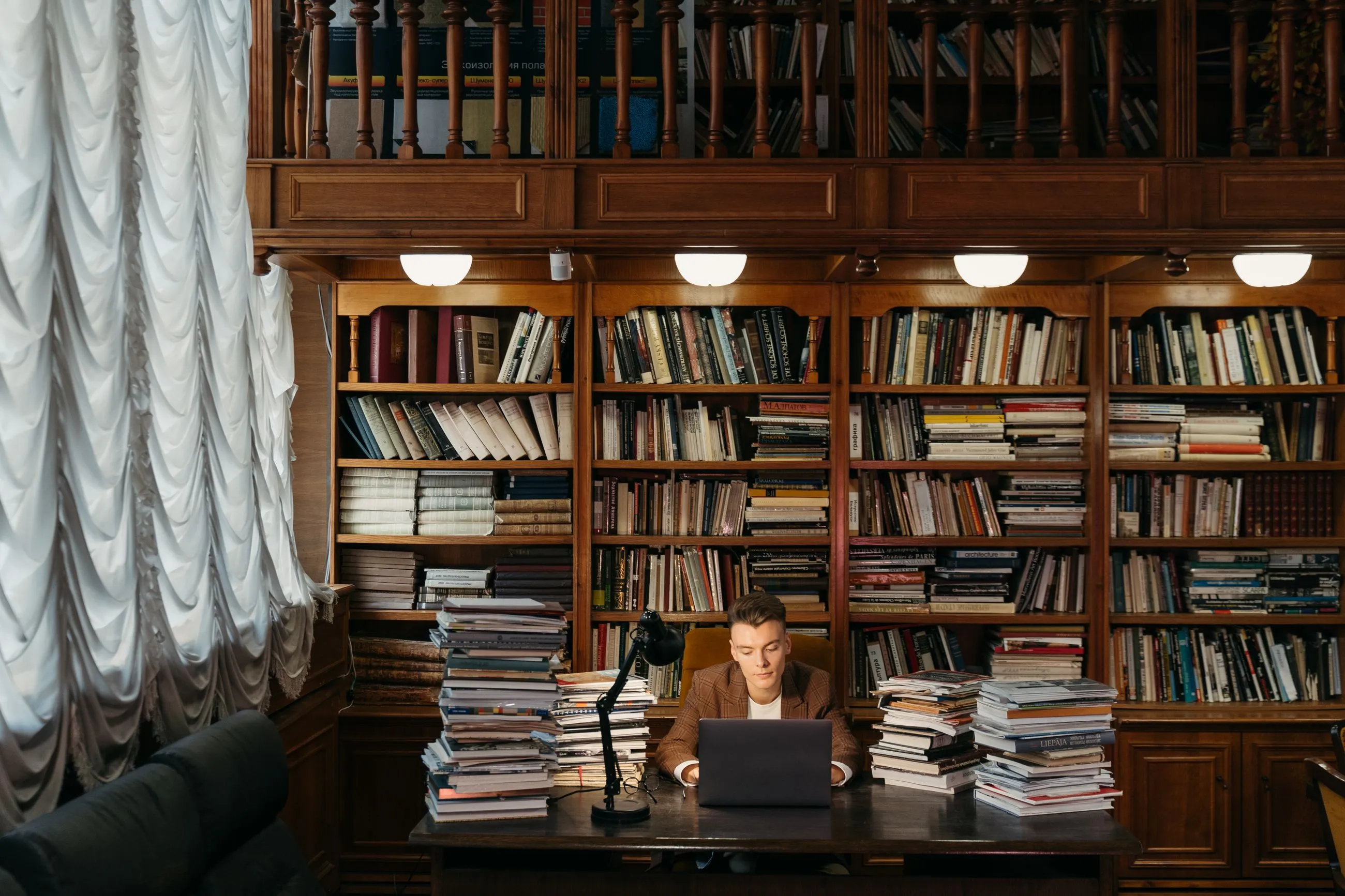 Joven estudiando con su portátil entre pilas de libros en una biblioteca clásica