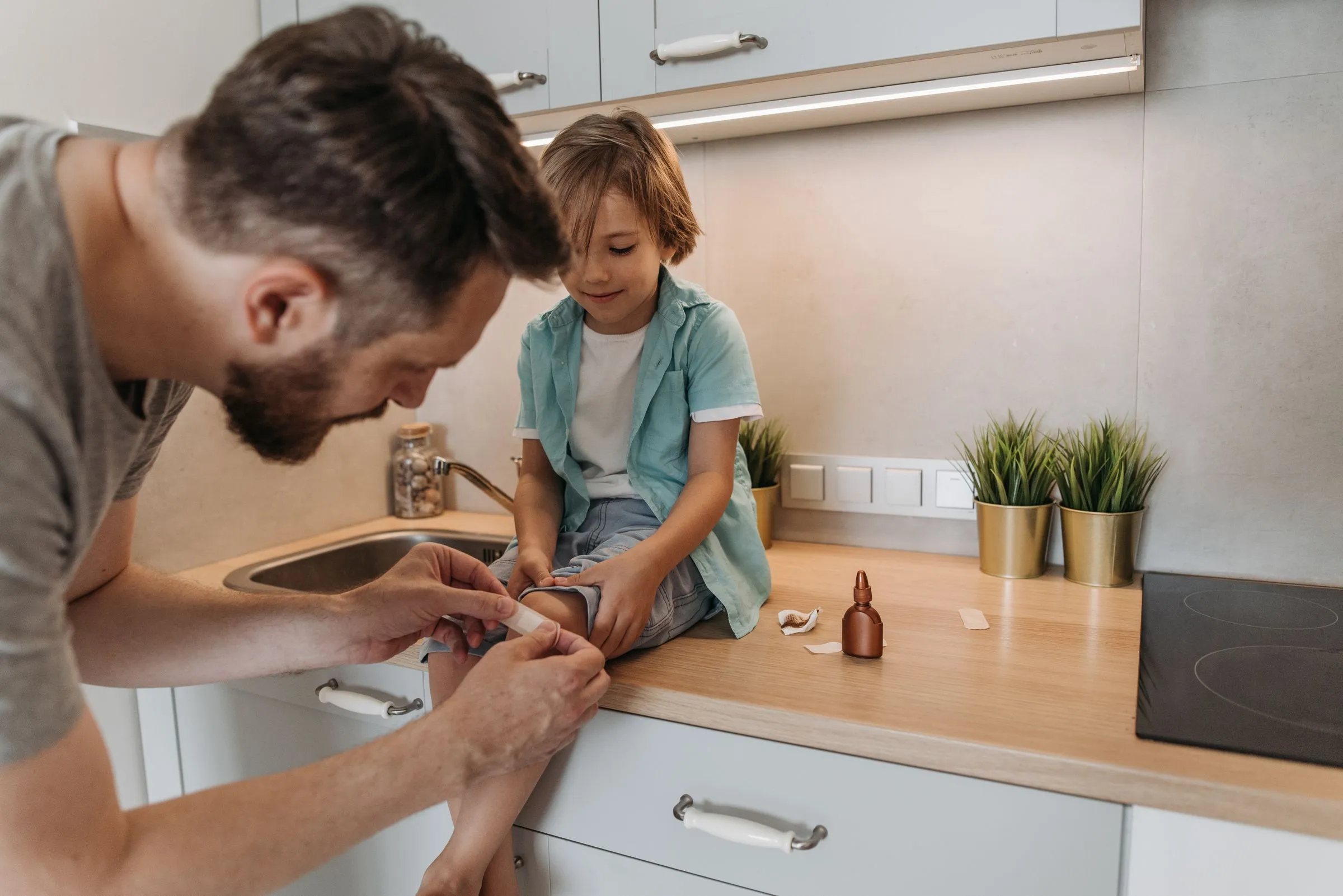 Un padre pone una tirita en la rodilla de su hijo, sentado en la encimera de la cocina