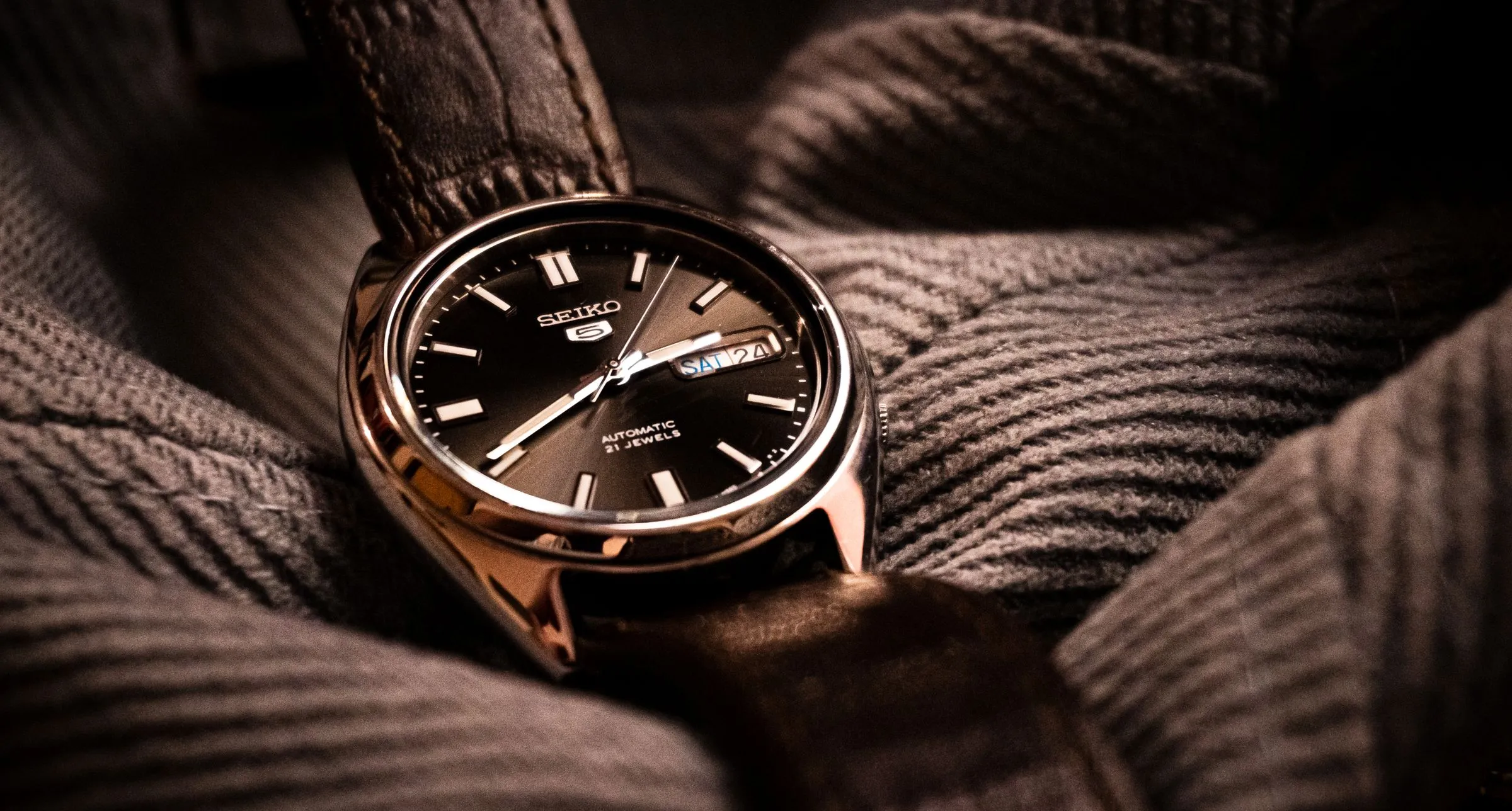 Seiko 5 clásico de esfera negra con calendario, posado sobre un jersey de punto gris